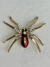 Gold spider-shaped brooch with red center and clear gemstones on a light gray background