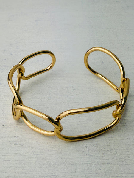 Modern gold-tone cuff bracelet with open link design, displayed on a table.