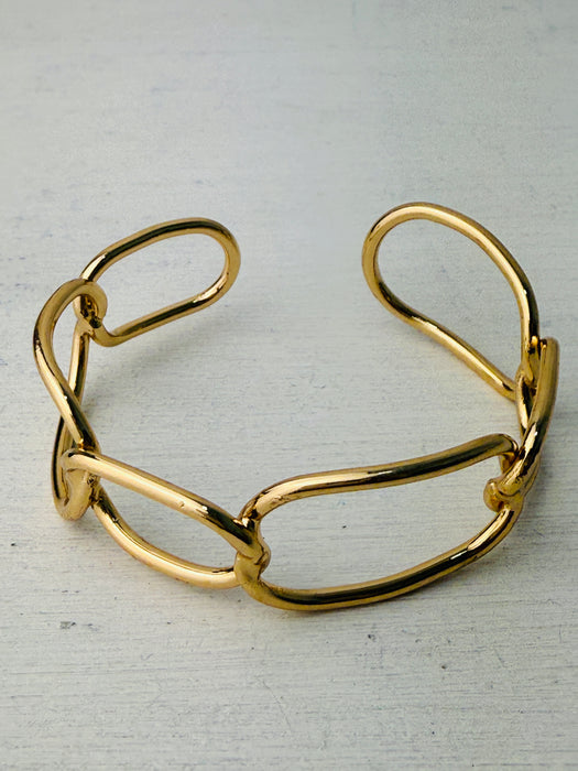 Modern gold-tone cuff bracelet with open link design, displayed on a table.