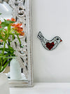 Mosaic glass bird with a heart on a wall, next to a decorative mirror and below a plant.