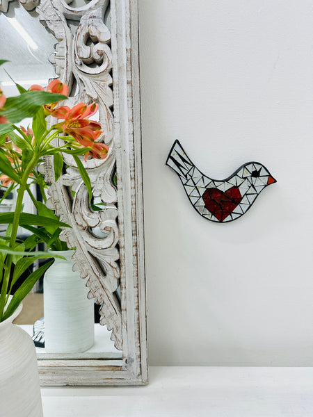 Mosaic glass bird with a heart on a wall, next to a decorative mirror and below a plant.