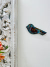 A hand painted glass mosaic in the shape of a blue bird with gold accents, mounted on a white wall next to a decorative floral panel.