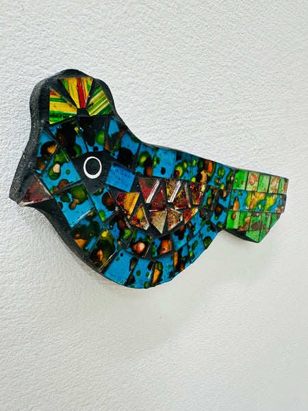 A hand painted glass mosaic in the shape of a blue and green bird with gold accents.