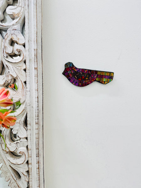 A hand painted glass mosaic in the shape of a violet bird with gold accents, mounted on a white wall next to a decorative frame.