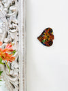 A hand painted glass mosaic in the shape of an angel fish with multicolored patterns and gold accents, mounted on a wooden surface next to a floral pattern.