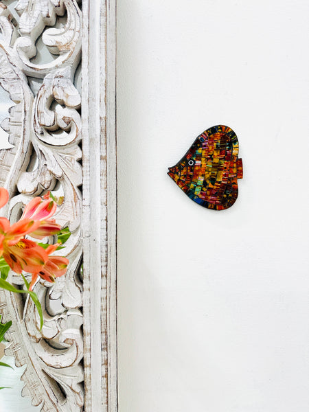 A hand painted glass mosaic in the shape of an angel fish with multicolored patterns and gold accents, mounted on a wooden surface next to a floral pattern.
