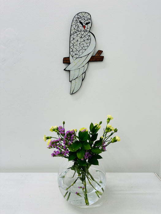 A handmade and hand painted mosaic glass owl mounted on a wall above a vase with flowers.