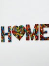 A mosaic wall piece with the word 'HOME' spelled out using multicolored hand painted glass tiles on a white background.