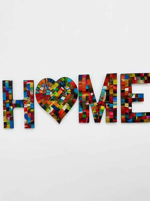 A mosaic wall piece with the word 'HOME' spelled out using multicolored hand painted glass tiles on a white background.