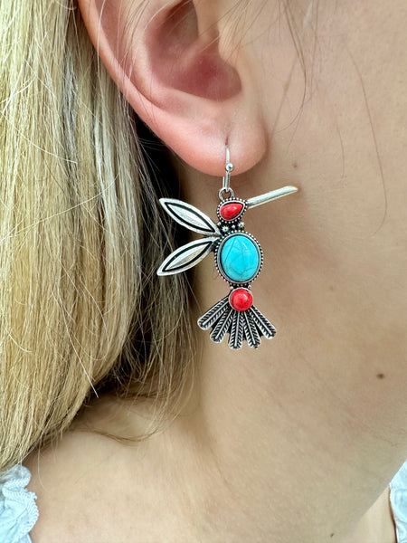 A person wearing a pair of silver-colored bird-shaped dangle earrings with a blue stone and red accents.