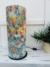 display view of mosaic cylinder lamp in multi with light off on a white surface next to a plant pot