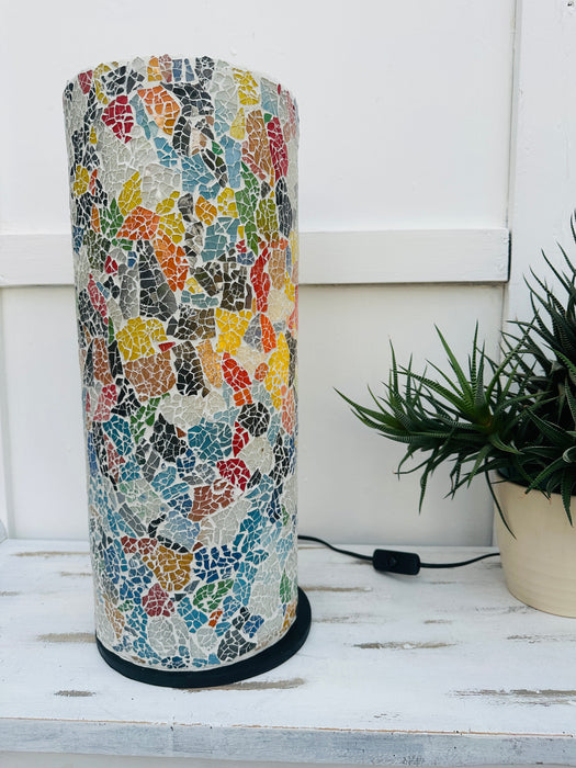 display view of mosaic cylinder lamp in multi with light off on a white surface next to a plant pot