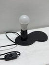 a black swivel socket holding a frosted white bulb, and a black power cord with a switch, set against a light gray wall and white surface.