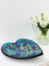 Handmade mosaic heart-shaped bowl in cyan tones on white table beside green flowers, showing glass mosaic pattern and black rim.