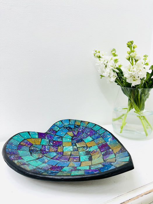 Handmade mosaic heart-shaped bowl in cyan tones on white table beside green flowers, showing glass mosaic pattern and black rim.
