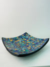 Angled view of handmade mosaic square plate showing terracotta base and glossy cyan glass surface on white background.