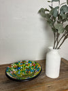 Handmade mosaic glass bowl in citrus colours placed on wooden table beside white vase with flowers.