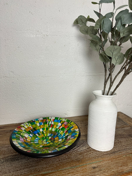 Handmade mosaic glass bowl in citrus colours placed on wooden table beside white vase with flowers.
