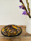 Handmade mosaic glass bowl in Siena tones on wooden table beside white vase with purple flower, showing warm golden mosaic design.