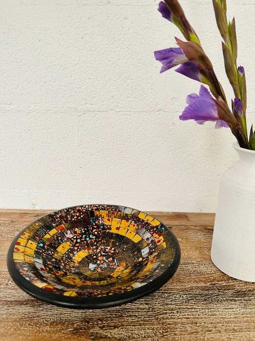 Handmade mosaic glass bowl in Siena tones on wooden table beside white vase with purple flower, showing warm golden mosaic design.