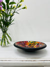 Handmade mosaic glass bowl in Calypso tones on white table beside green flowers, showing red, yellow and gold glass mosaic pattern with black rim.