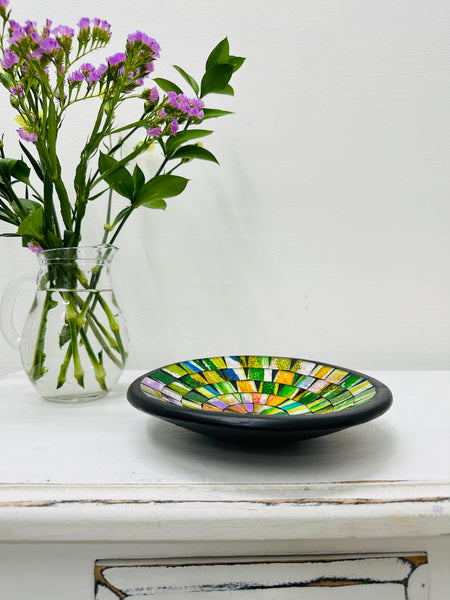 Handmade mosaic glass bowl in citrus tones on white table beside purple flowers, showing yellow and green glass pattern with black rim.