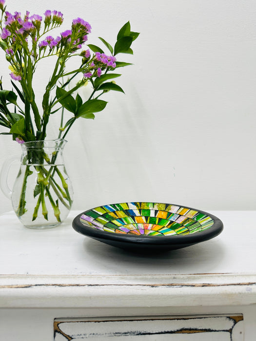 Handmade mosaic glass bowl in citrus tones on white table beside purple flowers, showing yellow and green glass pattern with black rim.