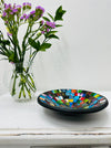 Handmade mosaic glass bowl in Sterope tones on white table beside purple flowers, showing colourful glass pattern with black rim.