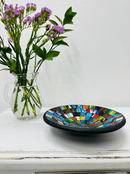 Handmade mosaic glass bowl in Sterope tones on white table beside purple flowers, showing colourful glass pattern with black rim.