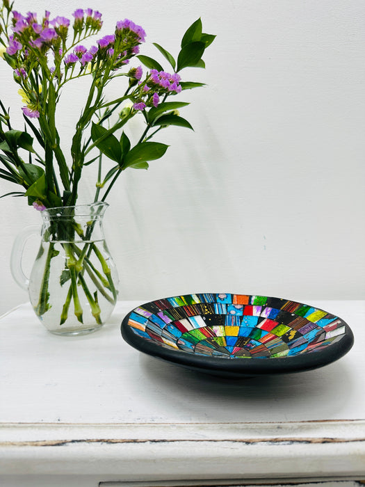 Handmade mosaic glass bowl in Sterope tones on white table beside purple flowers, showing colourful glass pattern with black rim.