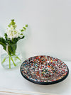 Handmade mosaic glass bowl in vibrant spectrum tones on white table beside green plant, showing colourful glass pattern and terracotta base.
