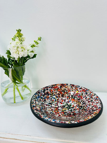 Handmade mosaic glass bowl in vibrant spectrum tones on white table beside green plant, showing colourful glass pattern and terracotta base.
