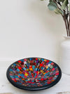 Handmade mosaic glass bowl in Starburst tones on white table beside vase with green leaves, showing bright red and gold mosaic surface.