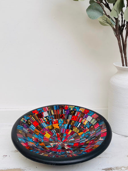 Handmade mosaic glass bowl in Starburst tones on white table beside vase with green leaves, showing bright red and gold mosaic surface.