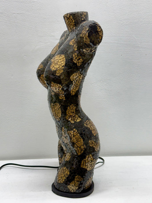 A sculptural female torso lamp or decorative figure with a glossy black and gold crackle mosaic pattern, standing on a black circular base against a light gray wall.