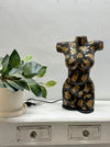 A decorative torso-shaped lamp or sculpture in glossy black with metallic gold floral patterns, displayed on a white cabinet next to a potted green plant against a light gray wall.