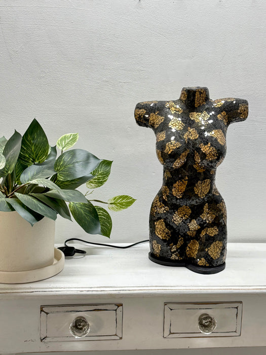 A decorative torso-shaped lamp or sculpture in glossy black with metallic gold floral patterns, displayed on a white cabinet next to a potted green plant against a light gray wall.