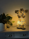 A decorative illuminated torso-shaped lamp with a gold and black floral pattern sits on a white dresser next to a potted plant, casting a warm yellow glow against a neutral wall.