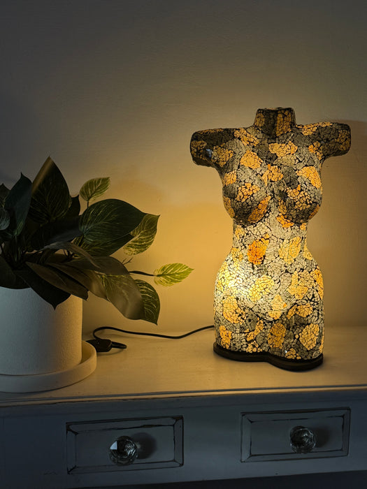 A decorative illuminated torso-shaped lamp with a gold and black floral pattern sits on a white dresser next to a potted plant, casting a warm yellow glow against a neutral wall.