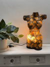 A decorative torso-shaped lamp with a glossy black and gold marbled finish, glowing warmly from within on a white console table next to a potted plant.