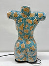 A ceramic female torso sculpture with a mosaic tile pattern in teal, gold, and white, standing on a black base against a light gray wall.