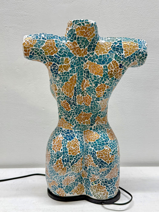 A ceramic female torso sculpture with a mosaic tile pattern in teal, gold, and white, standing on a black base against a light gray wall.