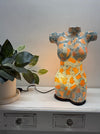 A decorative torso-shaped lamp with a mosaic pattern of gold, turquoise, sitting on a white wooden cabinet next to a potted plant.