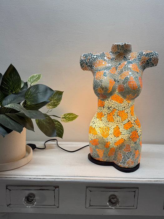A decorative torso-shaped lamp with a mosaic pattern of gold, turquoise, sitting on a white wooden cabinet next to a potted plant.