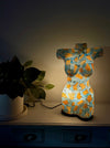A decorative torso-shaped lamp with a turquoise and gold textured pattern, glowing on a dark wooden cabinet next to a potted plant.