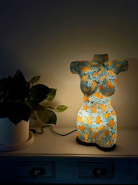 A decorative torso-shaped lamp with a turquoise and gold textured pattern, glowing on a dark wooden cabinet next to a potted plant.