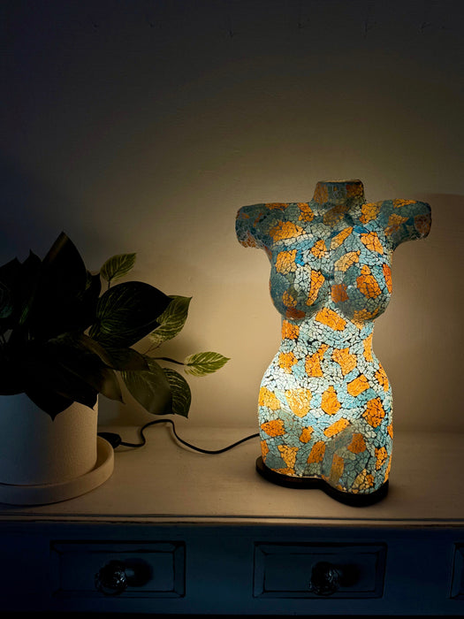 A decorative torso-shaped lamp with a turquoise and gold textured pattern, glowing on a dark wooden cabinet next to a potted plant.