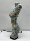 A sculptural, curved female-form lamp or decorative object covered in a mosaic tile pattern of teal blue, gold, and white, standing on a black circular base against a light gray wall.