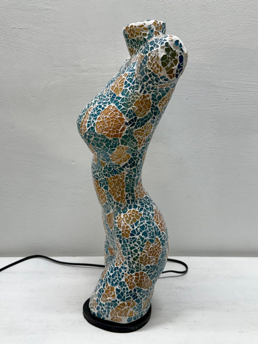 A sculptural, curved female-form lamp or decorative object covered in a mosaic tile pattern of teal blue, gold, and white, standing on a black circular base against a light gray wall.