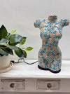 A ceramic torso-shaped lamp or figurine with a turquoise, white, and tan mosaic pattern sits on a white wooden cabinet next to a potted green plant.